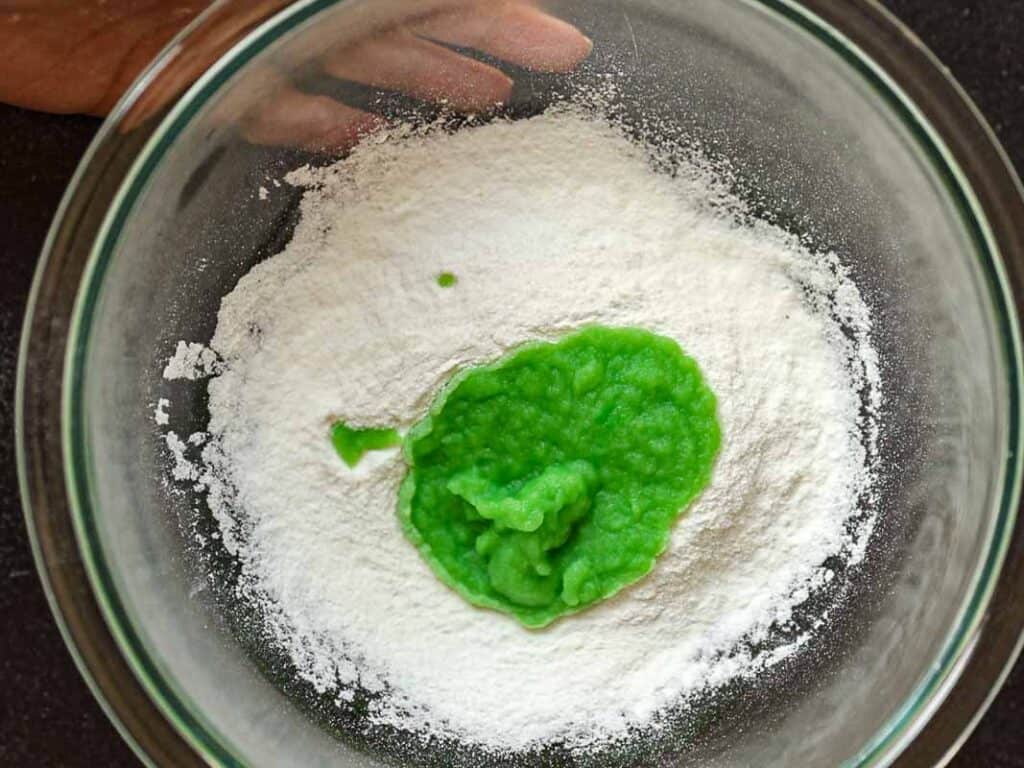 A glass bowl containing white rice flour and bright green puree on a dark surface. A hand is partially visible at the top edge of the bowl.